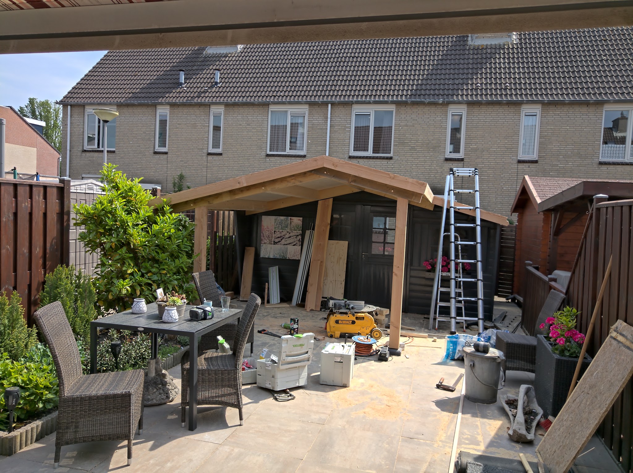 Houten overkapping in aanbouw op een terras, met tuinset, ladder en zaagtafel