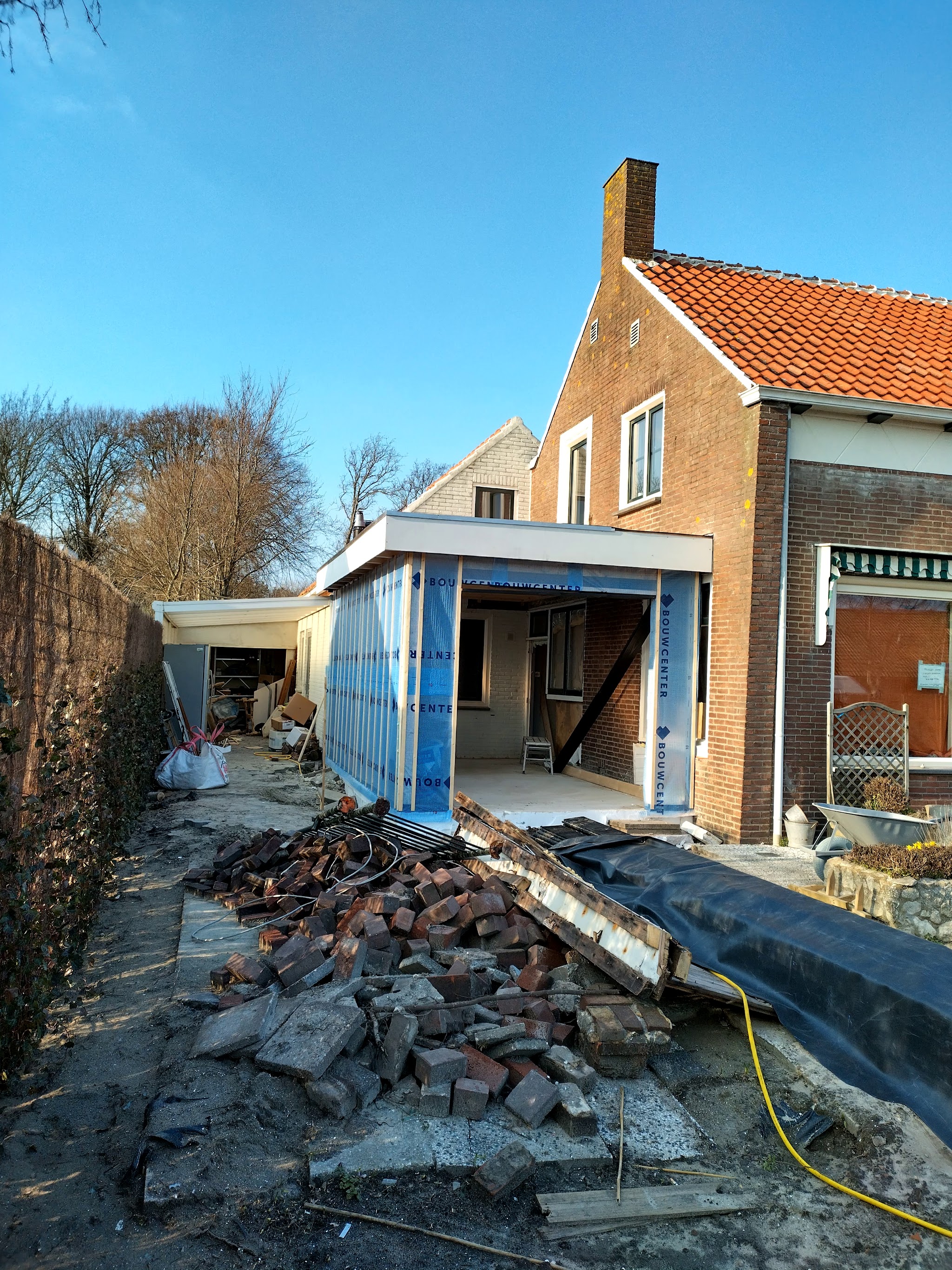 Voorbereiding van een aanbouw: gesloopte tuinmuur en puin naast de woning, met de nieuwe constructie in opbouw